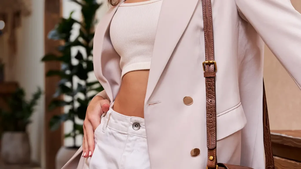 A woman wearing a light beige blazer over a white ribbed crop top and white high-waisted pants, posing with one hand on her hip and a brown handbag over her shoulder, set against a backdrop of indoor plants.