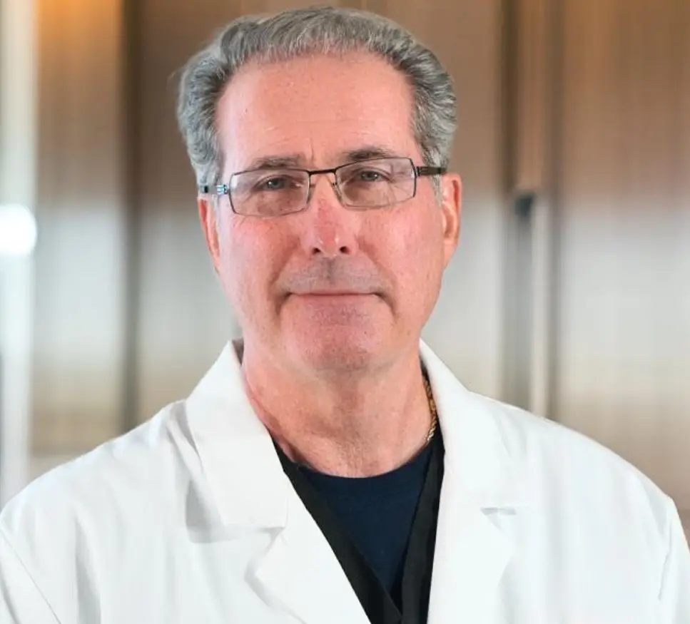 Portrait of a male medical professional wearing glasses and a white lab coat, standing in a clinical setting with a neutral background.