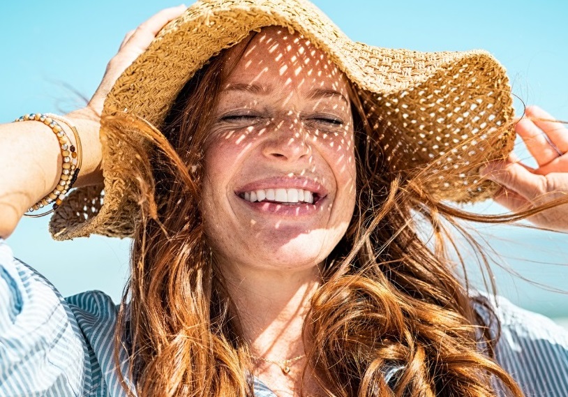 A woman with long, wavy red hair smiles brightly while wearing a wide-brimmed straw hat. Sunlight filters through the hat, casting playful shadows on her face. She is outdoors, with a clear blue sky in the background, suggesting a sunny day at the beach. Her joyful expression conveys a sense of happiness and relaxation, making it an ideal image for themes related to summer, leisure, and outdoor activities. The photo captures the essence of enjoying nature and the warmth of the sun.