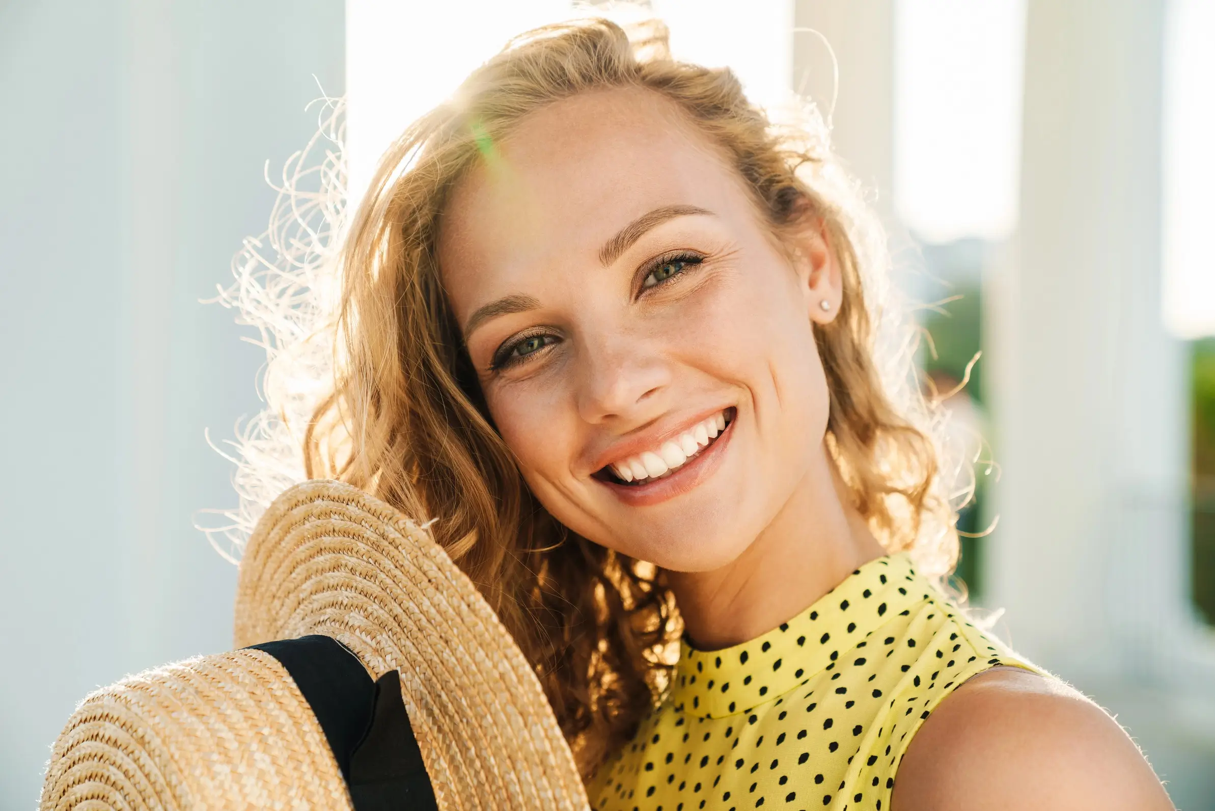 Smiling young woman with curly hair holding a straw hat, wearing a yellow polka dot top. The background features soft sunlight, enhancing her cheerful expression. This image conveys a sense of joy and summer vibes, making it ideal for lifestyle and fashion content. Perfect for articles or blogs about summer fashion, outdoor activities, or personal style tips.