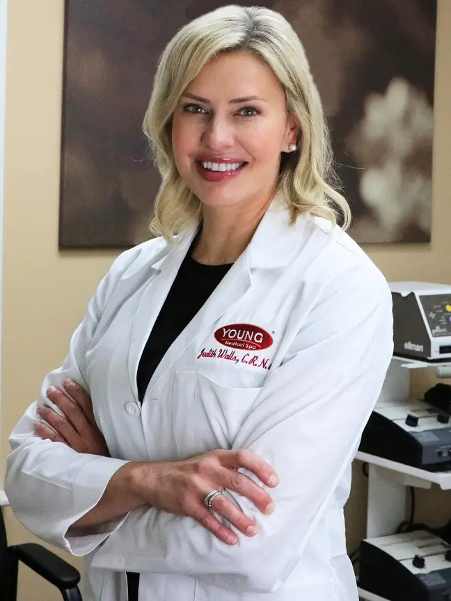 Professional female medical practitioner wearing a white coat with a name badge, smiling confidently with arms crossed. The background features medical equipment, indicating a clinical environment. This image represents expertise in medical aesthetics and patient care, highlighting a welcoming and professional atmosphere in a medical spa setting. Ideal for showcasing services related to skincare and wellness.