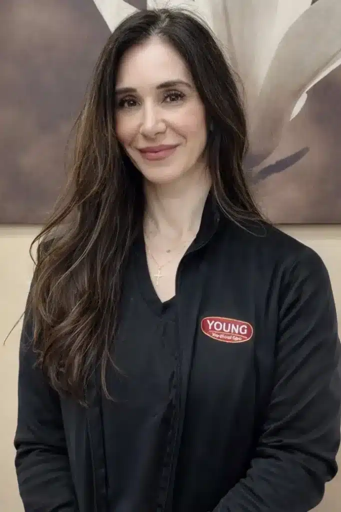 Portrait of a woman with long dark hair, wearing a black jacket with a "YOUNG" logo, standing in front of a neutral-toned background featuring a large floral design. She has a warm smile and is wearing a simple necklace. This image conveys a professional and friendly atmosphere, suitable for a business or wellness setting, such as a spa or aesthetic clinic. The woman appears approachable and confident, enhancing the welcoming vibe of the establishment.