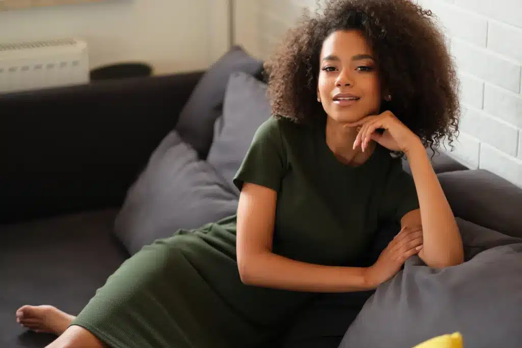 A woman with curly hair is sitting comfortably on a dark gray sofa, wearing a short-sleeved green dress. She rests her chin on her hand, smiling at the camera, conveying a relaxed and confident demeanor. The background features a light-colored brick wall and a window, creating a cozy atmosphere. This image represents themes of comfort, style, and modern living.